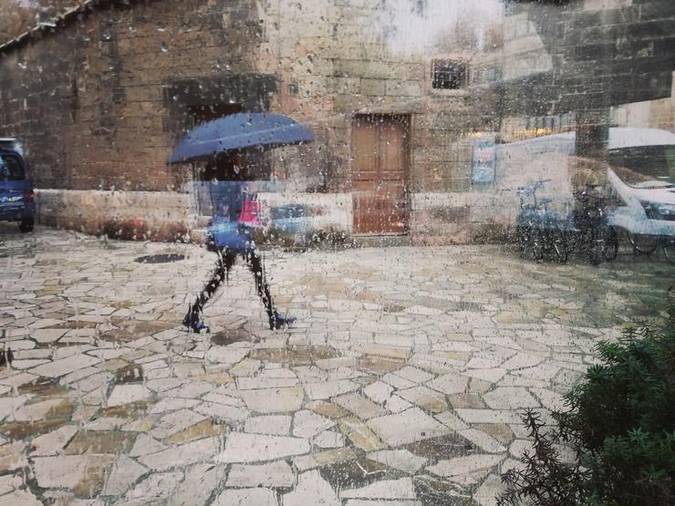 Unha muller pasea polo centro de Palma cun paraugas, vista a través dun cristal con pingas de choiva. 