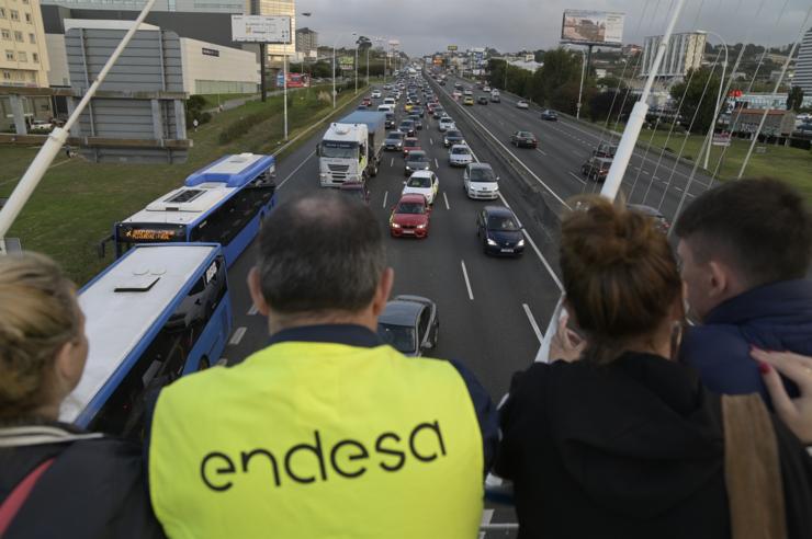 Varias persoas, unha cun chaleco de 'Endesa', observan unha caravana de automóbiles e camións mobilizada pola paralización da central térmica de Endesa en de As Pontes diríxese desde esa localidade coruñesa cara á Coruña en protesta polo parón,. M. Dylan - Europa Press 