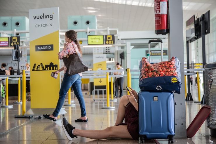 Un mozo mira o seu movil sentado xunto á súa maleta, xunto ao posto de Check-in da compañía, Vueling no Aeroporto 