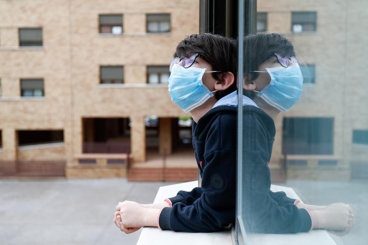 Un neno cunha máscara asómase á xanela da súa casa durante o confinamento polo coronavirus, en Valdemoro