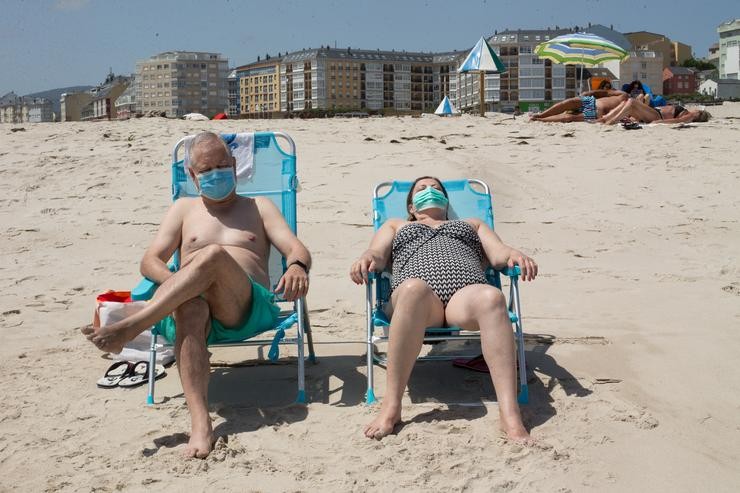 Dúas persoas protexidas con máscaras toman o sol na praia da Rapadoira en Foz, na comarca da Mariña, Lugo 