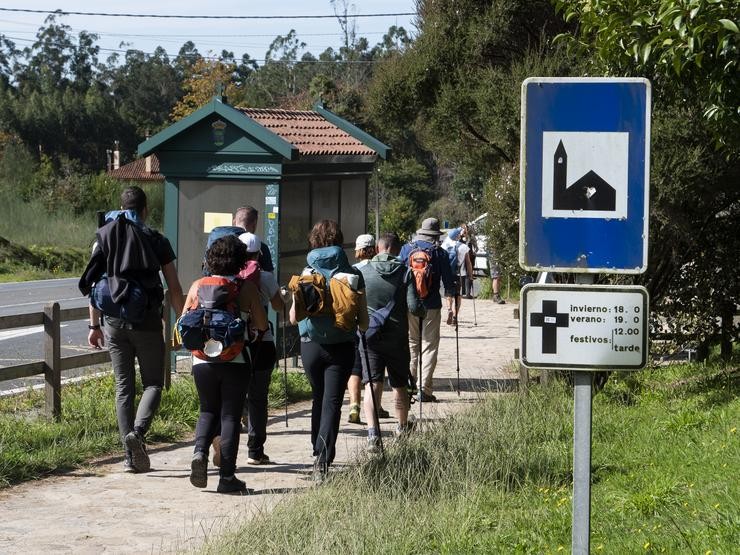 Un grupo de peregrinos pasa fronte a un cartel que indica o horario de misas mentres realizan o Camiño de Santiago / César Arxina